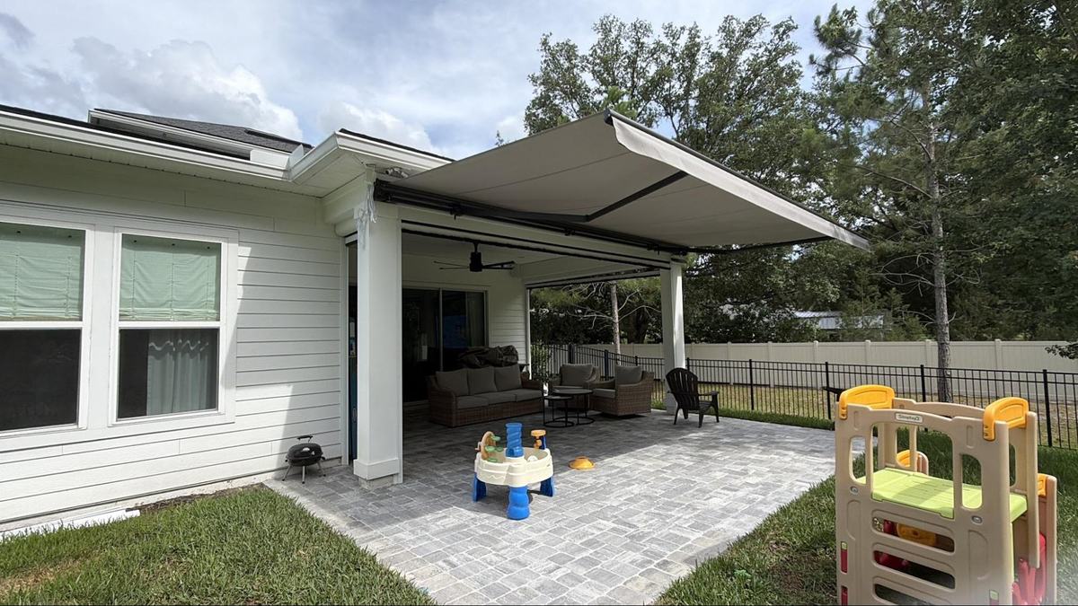 Beige retractable awning extended over a paver patio with children's play equipment and outdoor furniture at a Jacksonville area home