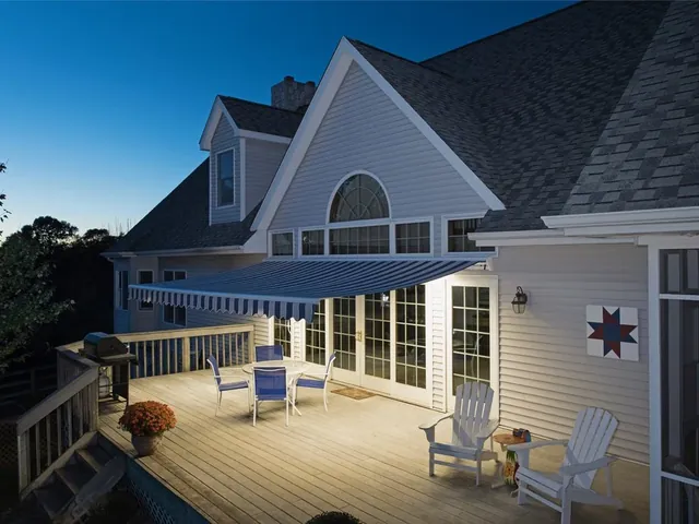 Motorized awning with integrated LED lighting over a gray lap-siding home in the evening